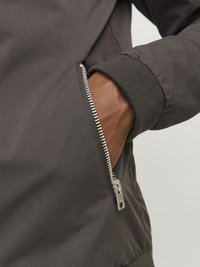 Gray jacket featuring a zippered pocket, ribbed cuff, and smooth fabric texture. The zipper is metallic, running parallel to the pocket opening.