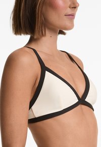 Top bikini triangolare crema con bordi neri, dotato di spalline sottili e design incrociato sul davanti. Texture liscia.