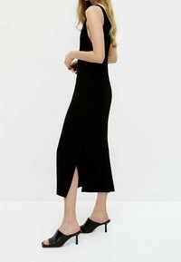 Schwarzes, geripptes Strickkleid mit körperbetontem Schnitt, seitlichem Schlitz und ärmellosem Design, kombiniert mit schwarzen, hohen Mules. Schlichte und elegante Silhouette.