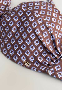 Tissu marron avec motif losanges bleu et blanc, froncé sur le bord avec un petit charme rond doré attaché.