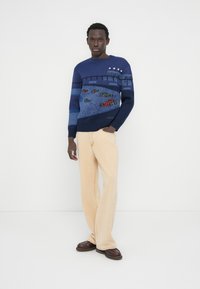 Πλεκτό πουλόβερ σε ναυτικό μπλε χρώμα με σχέδιο αγωνιστικού αυτοκινήτου, συνδυασμένο με ανοιχτό μπεζ παντελόνι καμπάνα και καφέ παπούτσια.