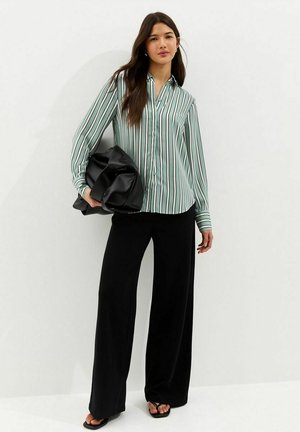 Kobieta w zielono-białej paskowanej bluzce i czarnych luźnych spodniach, trzymająca dużą czarną torbę, stojąca na jednolitym białym tle.