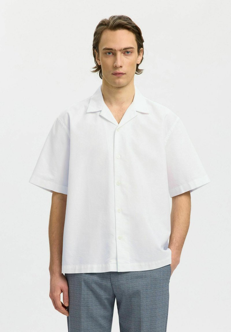 Jeune homme aux cheveux bruns mi-longs portant une chemise blanche à manches courtes boutonnée et un pantalon gris, debout avec une main dans la poche.