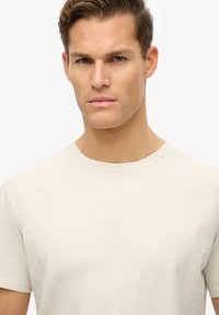 Beige bomulds t-shirt med korte ærmer og rund halsudskæring, der har et subtilt logo på brystet og en blød, glat tekstur.