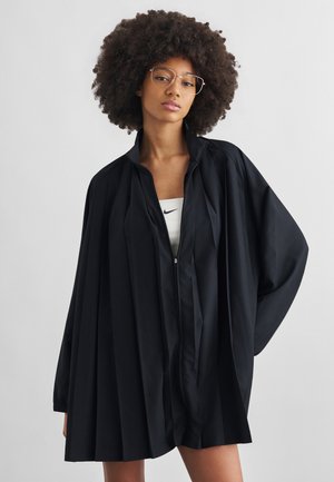 Moteris su garbanotais plaukais ir akiniais, dėvinti neregią juodą klostuotą zip-up striukę ant baltos Nike palaidinės vientiso fono.