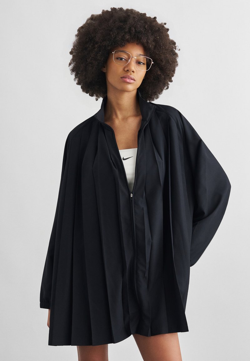 Frau mit lockigem Haar und Brille, die eine lockere schwarze, plissierte Jacke mit Reißverschluss über einem weißen Nike-Oberteil vor einfachem Hintergrund trägt.