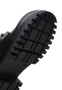 Suola di stivali in gomma nera con scanalature profonde e rigate. Il materiale sembra strutturato e resistente, progettato per garantire aderenza su varie superfici.