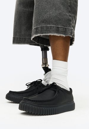 Onderbenen met zwarte sneakers, witte sokken en grijze denim shorts, met een prothetisch been onder de knie zichtbaar.
