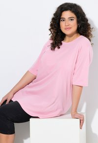 Helles rosa Übergrößen T-Shirt mit kurzen Ärmeln, aus weichem Baumwollstoff. Verfügt über einen runden Ausschnitt und eine lockere Silhouette, kombiniert mit schwarzen Shorts.