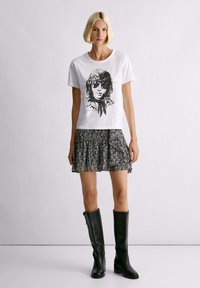 Witte katoenen t-shirt met een zwarte grafische print van een gezicht; gedragen met een geplooide zwart-wit gestreepte rok en kniehoog zwarte leren laarzen.