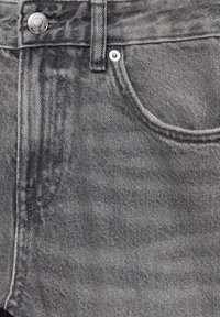 Gros plan d'une poche de jean en denim gris avec rivets métalliques, passants de ceinture et détails de couture visibles.