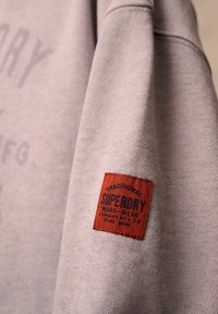 Felpa grigia con tessuto testurizzato, con un'etichetta ricamata rossa che mostra "SUPERDRY" e ulteriori dettagli in blu scuro.