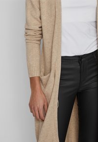 Beige stickad cardigan med ribbade manschetter och sidofickor, i kombination med svarta tajta byxor. Tyget ser mjukt och strukturerat ut.