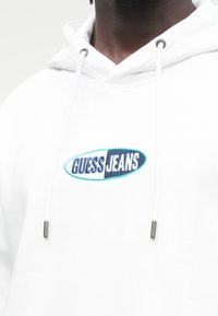Close-up de uma pessoa a usar um capucho branco com o logótipo "GUESS JEANS" bordado no peito e cordões brancos com pontas metálicas.