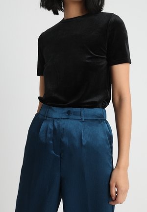 Czarna aksamitna koszulka z okrągłym dekoltem i krótkimi rękawami, zestawiona z turkusowymi satynowymi plisowanymi spodniami z zapięciem na guzik.