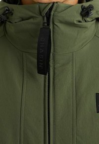 Olivově zelená bunda s kapucí, hladkým povrchem, předním zipem, detailem na šňůrky a černou nášivkou na hrudi.