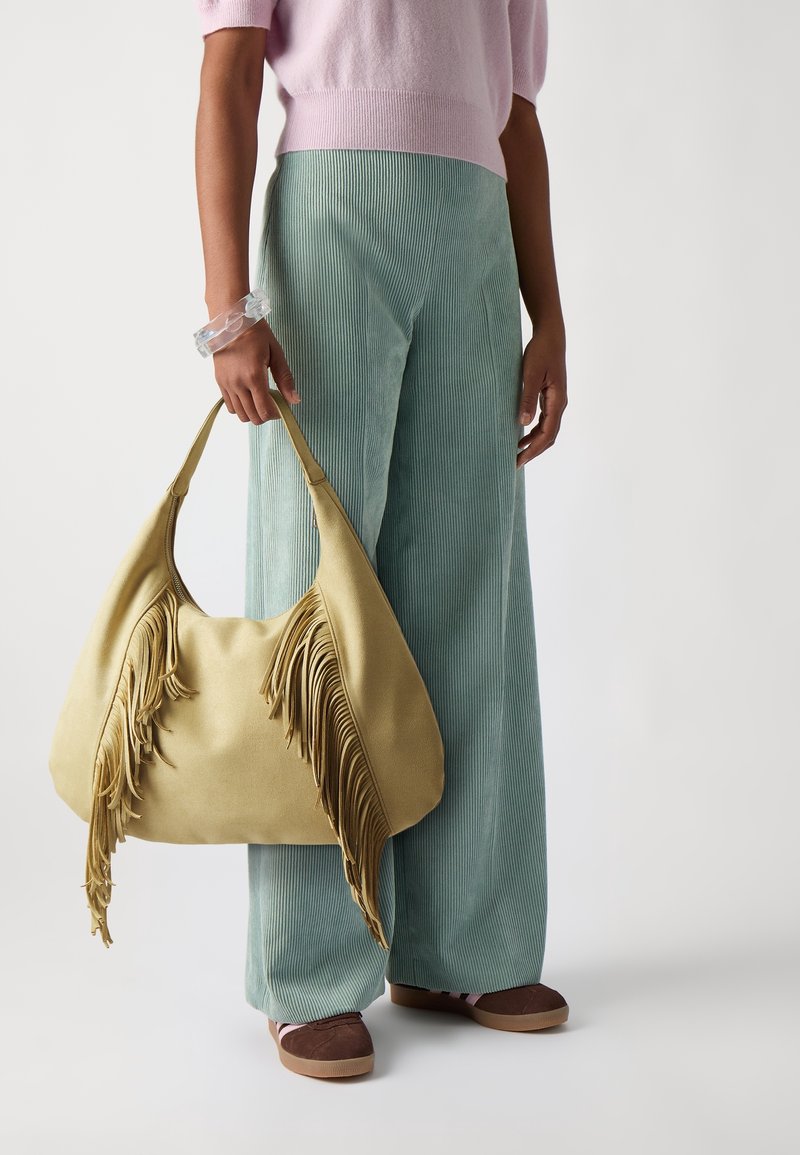 Sac à main à franges en matière jaune douce, avec une forme décontractée. Porté avec un pantalon large en velours côtelé vert clair et des chaussures marron à enfiler.