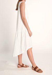 Weißes ärmelloses Kleid mit Stufen-Saum und asymmetrischem Schnitt. Kombiniert mit braunen flachen Sandalen mit Musterdesign. Weiche Textur.