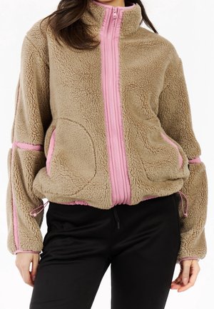 Femme portant une veste polaire beige avec une fermeture éclair et des bordures roses, associée à un pantalon noir, debout devant un fond blanc.