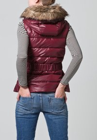 Smanicato imbottito bordeaux con cappuccio foderato in pelliccia, vita aderente e tessuto lucido, indossato sopra una maglia a maniche lunghe a righe bianco e nero e jeans blu.