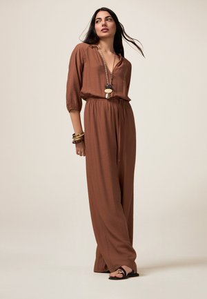 Brown, lightweight jumpsuit with a loose fit, three-quarter sleeves, adjustable waist, and full-length legs. Accessories include bracelets and a necklace.