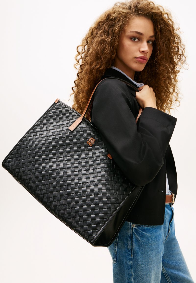 Νέα γυναίκα με σγουρά μαλλιά, φορώντας μαύρο μπουφάν και τζιν, κρατώντας μεγάλη μαύρη υφαντή τσάντα ώμου με καφέ λαβές.