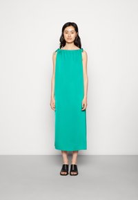 Um vestido maxi sem mangas em azul-turquesa, com detalhes de laço nos ombros, feito de um tecido suave. Usado com sandálias pretas de deslizar.