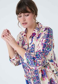 Long-sleeve blouse featuring a multicolored floral and patterned design on a beige background, with a button-up front and collar.