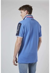 Chemise polo bleue avec un col rayé contrasté et des accents de manches sombres comportant des lettres blanches. Fabriquée en coton texturé.