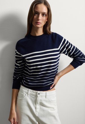 Marineblauwe katoenen trui met horizontale witte strepen, ronde hals en geribbelde mouwen. Gecombineerd met witte jeans staat het model tegen een lichte achtergrond.