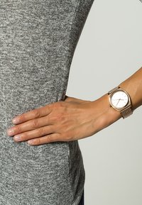 Roségoldene Armbanduhr mit minimalistischem weißen Zifferblatt, Metall-Mesh-Armband und Zeigern, die auf einer grauen strukturierten Oberfläche positioniert sind. Die Hand ruht auf der Taille.