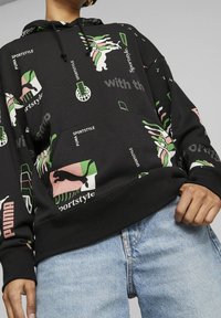 Sudadera negra con coloridos estampados gráficos de manos, logotipos y texto. El material parece suave; el diseño incluye una capucha y un bolsillo delantero.