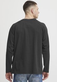 Black long-sleeve shirt with a round neckline, featuring a subtle logo detail on the back and a relaxed fit, paired with light blue jeans.