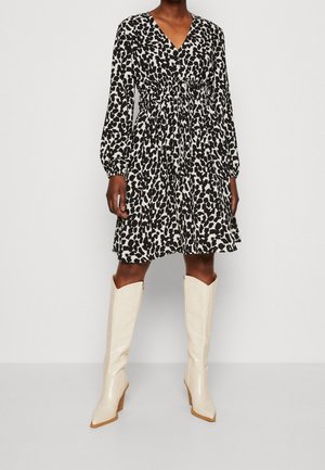 Vrouw draagt een zwart-witte dierenprint jurk met lange mouwen en een getailleerde taille, gecombineerd met witte kniehoge laarzen met hak, op een effen achtergrond.