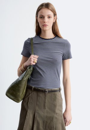 Jeune femme debout, visage tourné vers l’avant, portant un t-shirt rayé bleu, une jupe olive, des bijoux dorés et tenant un sac à main vert olive sur son épaule.