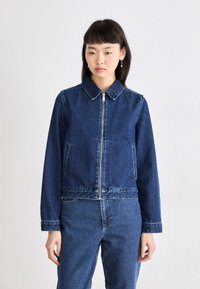 Denim striukė su užtrauktuku priekyje, smailiu apykakle, ilgomis rankovėmis ir šoninėmis kišenėmis; tamsiai mėlynos spalvos su šiek tiek išblukusia tekstūra.