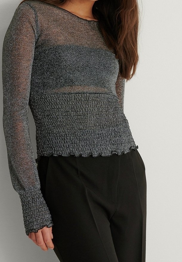 Femme portant un haut gris ajusté à manches longues, semi-transparent et scintillant, avec un tissu texturé aux poignets et à l'ourlet, associé à un pantalon noir taille haute.