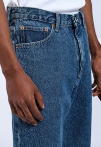 Jeans de mezclilla azul con un corte recto, remaches de cobre y un pequeño bolsillo delantero. La tela tiene una apariencia texturizada y ligeramente desgastada.