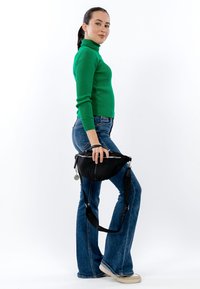 Černá textilní taška s zaobleným tvarem a stříbrným zipem, držená osobou v zeleném žebrovaném svetru a modrých rozšířených džínách, stojící bokem.