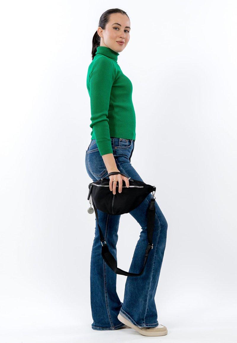 Černá textilní taška s zaobleným tvarem a stříbrným zipem, držená osobou v zeleném žebrovaném svetru a modrých rozšířených džínách, stojící bokem.