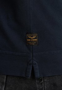 Granatowa teksturowana koszula z prostokątną metką z napisem "PME LEGEND" w złotym kolorze, z emblematem w skrzydłami i detalami wentylacyjnymi po bokach.