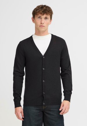 Black knit cardigan with a V-neckline, long sleeves, and front buttons. Ribbed cuffs and hem; worn over a white shirt.