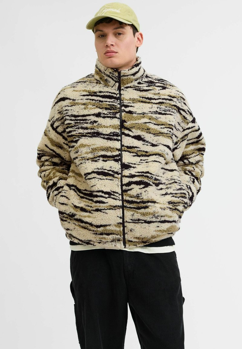 Chamarra de forro polar con cremallera en beige con rayas de tigre en negro y oliva, cuello alto, bolsillos frontales, usada sobre pantalones de pana negra.