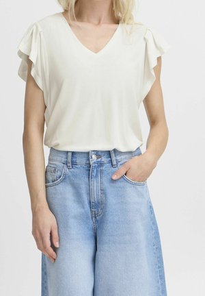Vrouw draagt een witte korte mouw V-hals blouse met ruches aan de mouwen en lichtblauwe high waist wijde jeans. Eén hand in de zak.