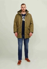 Mann trägt eine olivgrüne Kapuzenparka, dunkle jeans, braune Lederstiefel und ein navyblaues T-Shirt mit Grafik.
