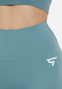Leggings de fitness de cintura alta en un suave color azul verdoso. Fabricados con una tela elástica, presentan un logo blanco en la cadera y costuras planas.