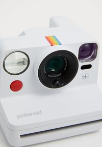 White instant camera with a black lens, flash, red button, and rainbow stripe. Features a Polaroid logo and a compact design.