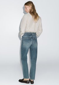 Hellblaue, hoch taillierte Jeans mit geradem Bein, aus ausgewaschenem Stretch-Denim und zwei Gesäßtaschen. Kombiniert mit einer gestreiften Bluse mit langem Arm.