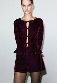 Woman wearing a dark red velvet long-sleeve top with front tie closures and matching shorts, standing against a plain light background.