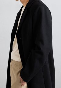 Abrigo de lana negro con un diseño estructurado, mangas largas y un solo botón, llevado sobre un suéter color crema y pantalones beige. Tejido texturizado visible.